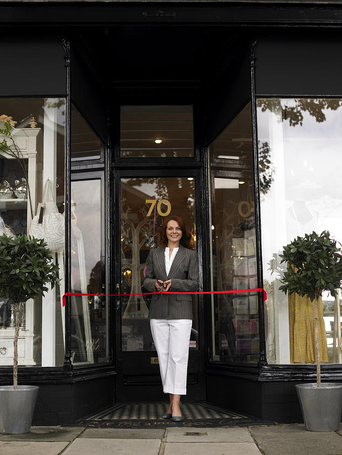 Young woman standing in entrance of new shop behind red ribbon, smiling, portrait Photograph by RL Productions