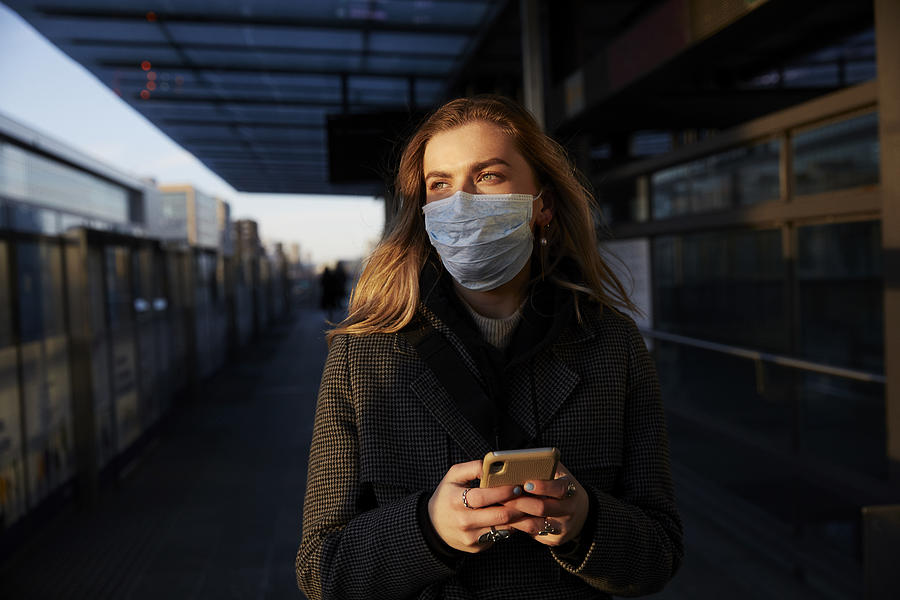 Young woman standing on train station wearing protective mask, using phone Photograph by Thomas Tolstrup