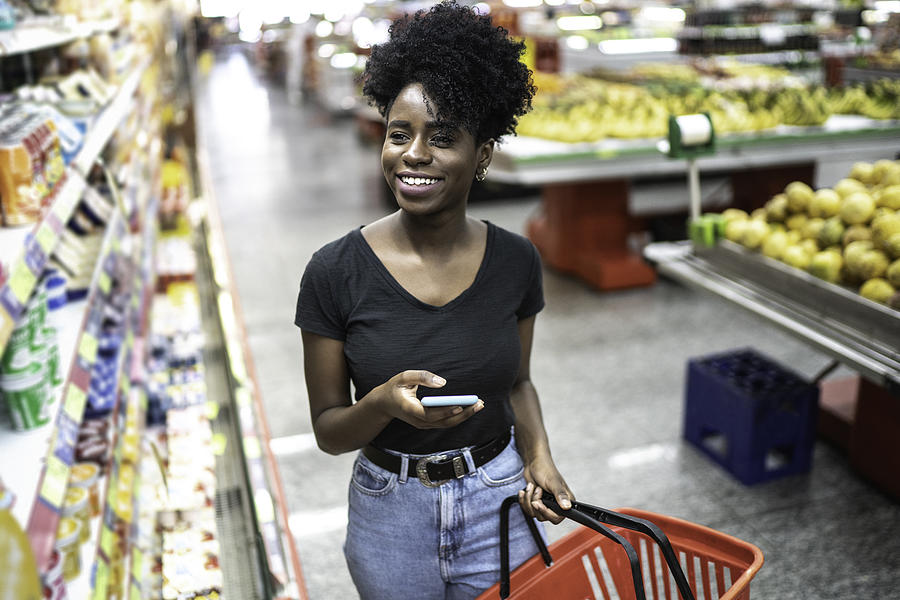 Young woman using mobile phone and choosing product in supermarket Photograph by FG Trade