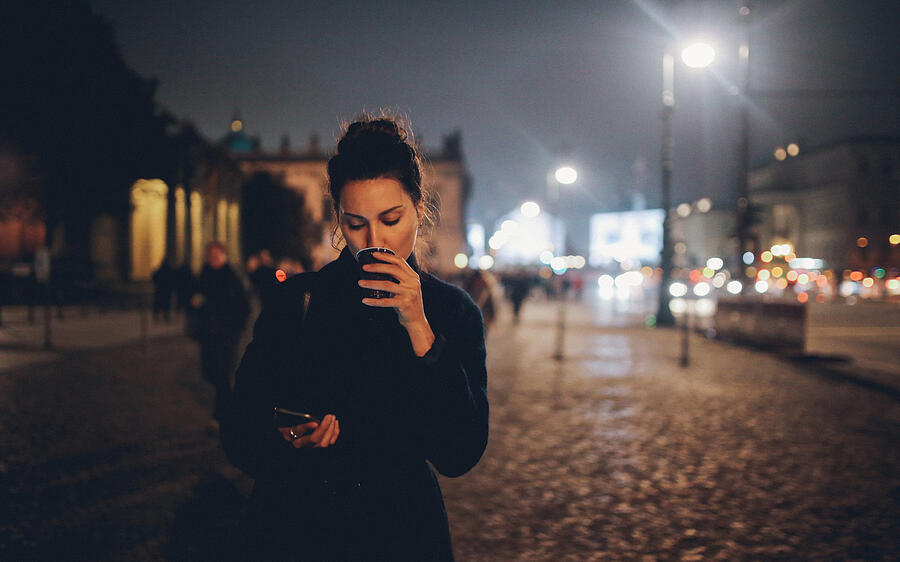 Young woman walking in Berlin at night checking phone Photograph by Lechatnoir