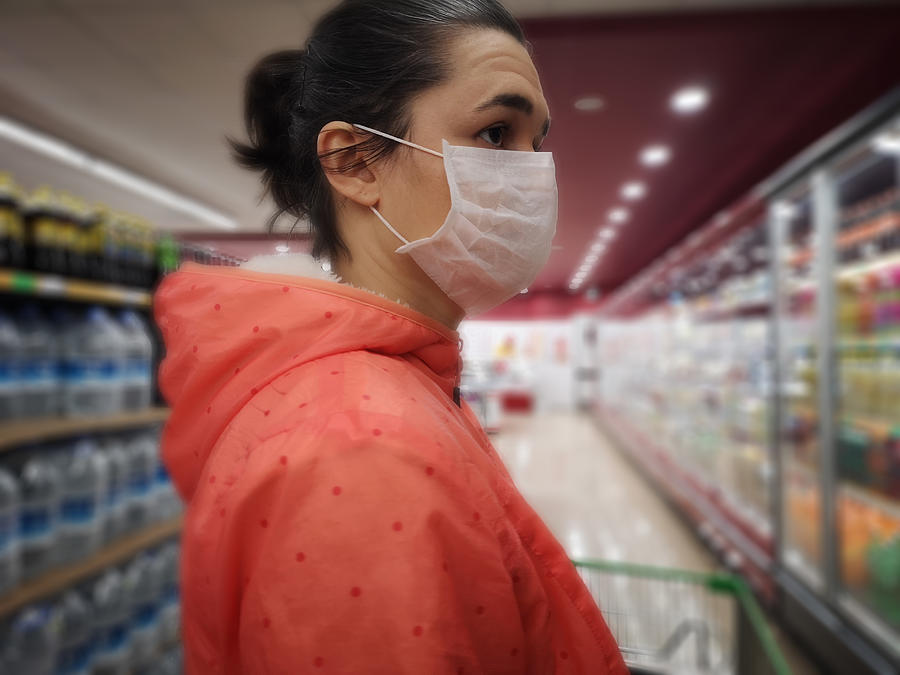 Young woman wears medical mask against virus while grocery shopping in supermarket, Photograph by Oonal