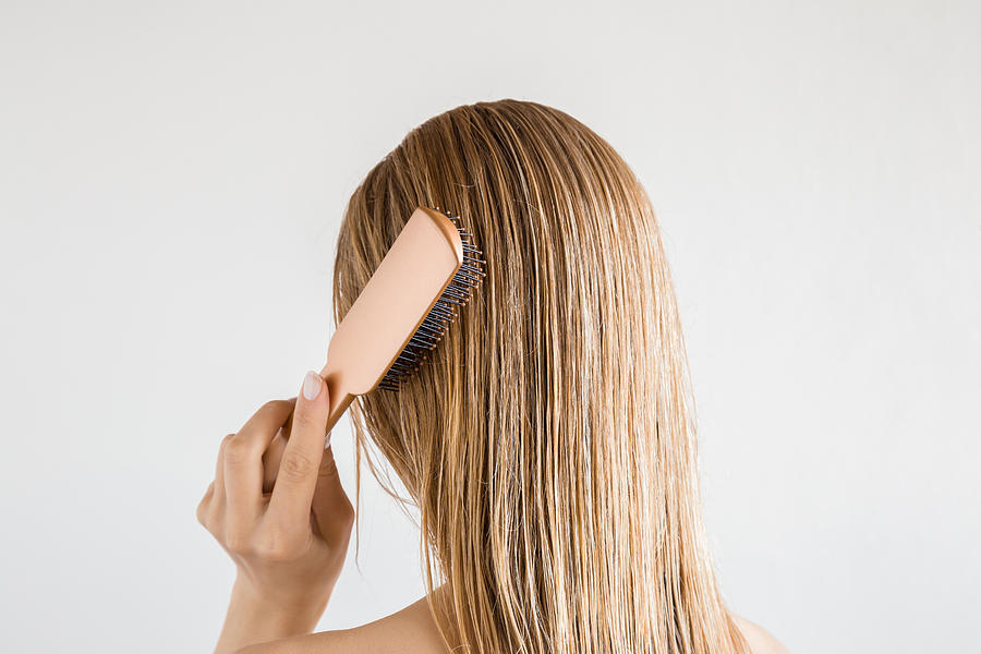 Young woman with comb brushing her wet, blonde, perfect hair after shower on the gray background. Care about beautiful, healthy and clean hair. Beauty salon concept. Back view. Photograph by FotoDuets